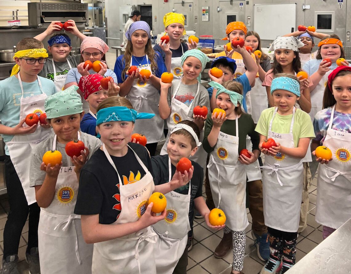 A group of kids in bandanas and aprons hold up tomatoes